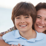Brunette boy with braces smiles on the beach with his brunette mother
